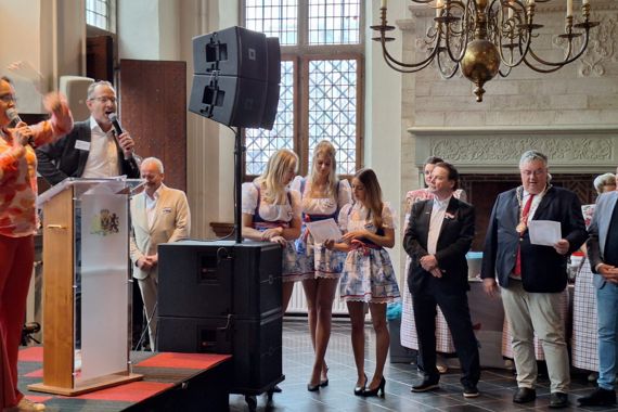 Links zingt Miel een lied achter een rostrum. rechts lezen verschillende mensen (waaronder burgemeester Bruls) de tekst mee.