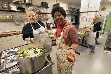 2 vrouwen staan voor het fornuis. Ze kijken in een grote pan met groente. Op de achtergrond snijdt een derde vrouw groente. Ze hebben alledrie een keukenschort aan.