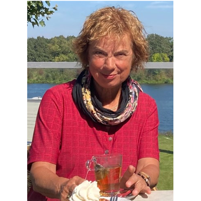 Gerrie zit aan een tafel met een kopje thee en een stuk taart. Ze heeft een rode blouse aan.  Op de achtergrond zie je een bos en een meer.