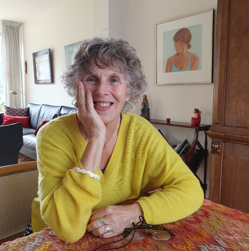 Marleen draagt een gele trui. Ze zit acher een tafel met een gekleurd tafellaken. Op de tafel ligt een bril. Marleen heeft een hand onder haar hoofd en lacht in de camera. Op de achtergrond is een woonkamer te zien.