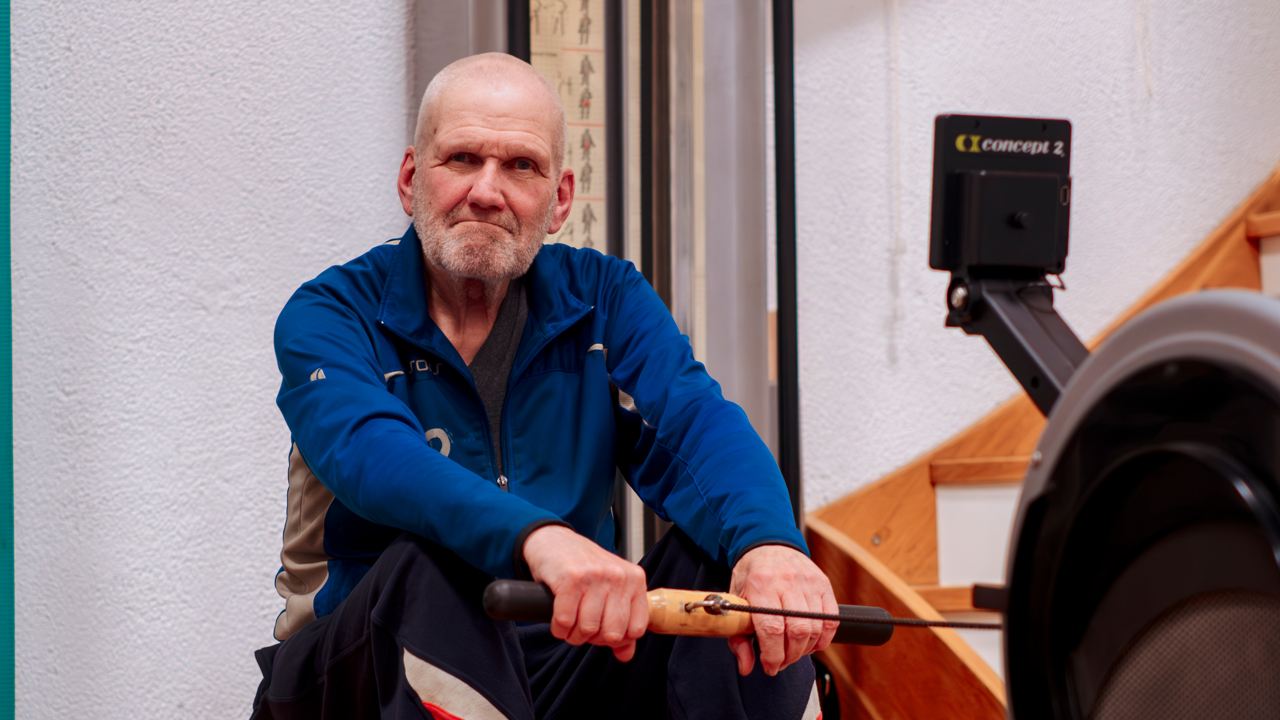 Piet zit op een roeimachine. Hij draagt een blauw sportvest. Hij kijkt tevreden in de camera.