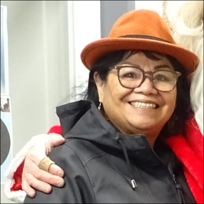 Vicky draagt een bruine bril, een oranje hoed een een zwart vest. Om haar schouder ligt de arm van de kerstman.
