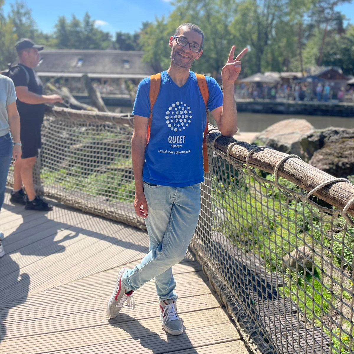 Arif maakt een peaceteken naar de camera. Hij leunt op het hek van een dierentuinverblijf. Hij draagt zijn Quietshirt, een lichte spijkerbroek en een oranje rugzak. op de achtergrond is een dierenverblijf te zien.