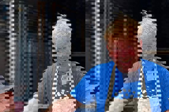 Hans draagt een Quiet shirt en een wit kookschort. Hij heeft zijn hand op de schouder van Jeroen en wijst met zijn andere hand naar hem. Hij lacht.