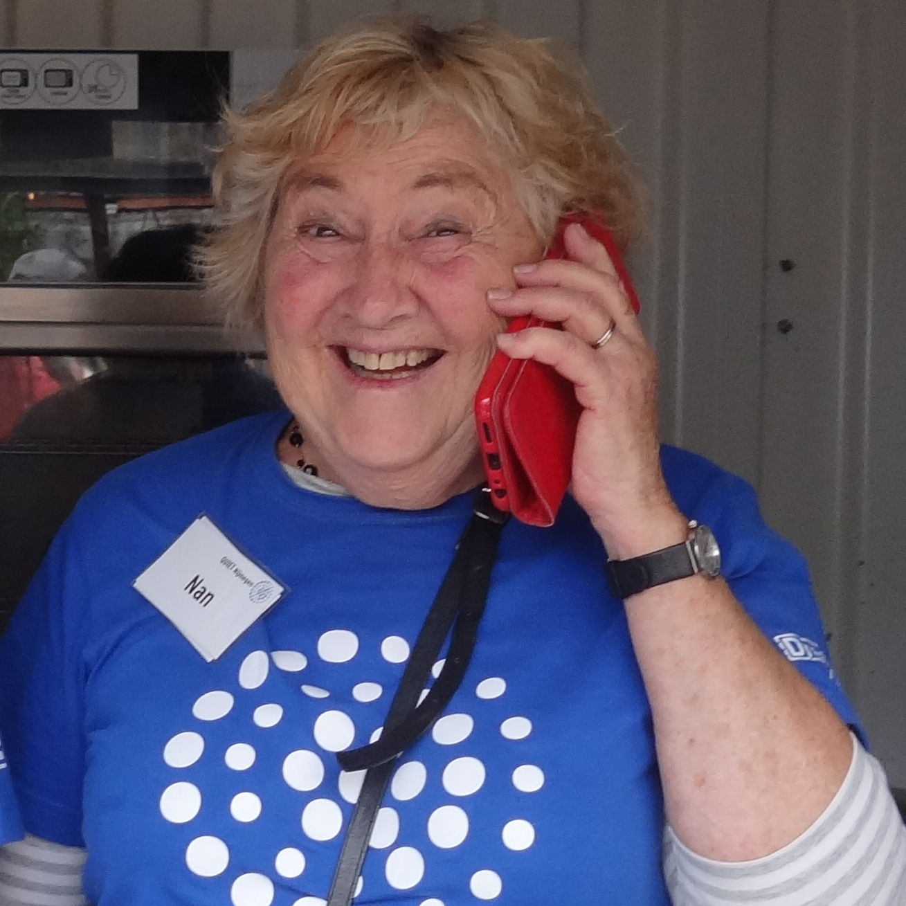 Nan heeft haar Quiet shirt aan. Ze houdt een rode mobiele telefoon tegen haar oor. Op de achtergrond zie je een klein stukje van de tot keuken verbouwde container waarin ze staat.