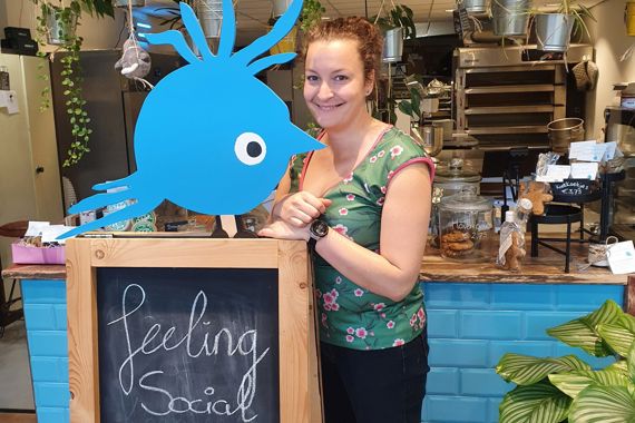 Chris leunt tegen het stoepbord van Koekkoek met de tekst "feeling social today?". Op de achtergrond zie je haar bakkerij.