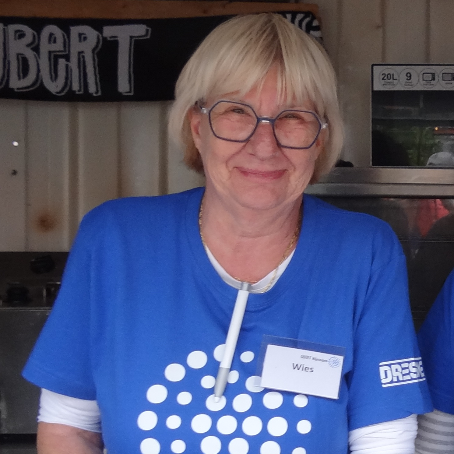 Wies heeft haar Quietshirt aan. Ze draagt een zeshoekige bril. Ze lacht in de camera. Op de achtergrond zie je een klein stukje van de omgebouwde container waarin ze staat.