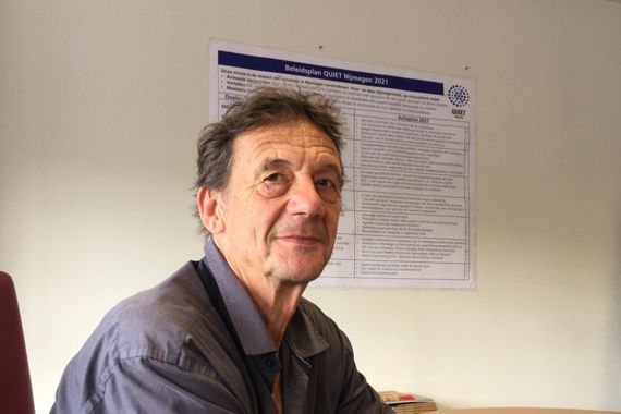 Henks handen liggen op de tafel voor hem. Hij kijkt sympathiek in de camera. Henk draagt een blauwe blouse en een grijze broek.