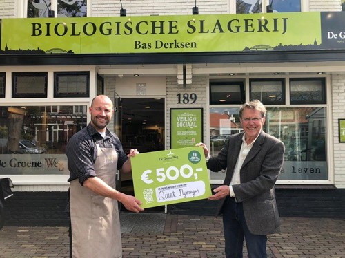 Vincent van Quiet Nijmegen en Bas Derksen poseren voor de gevel van de Biologische Slagerij van Bas. Ze hebben een bord vast met een donatie van €500.