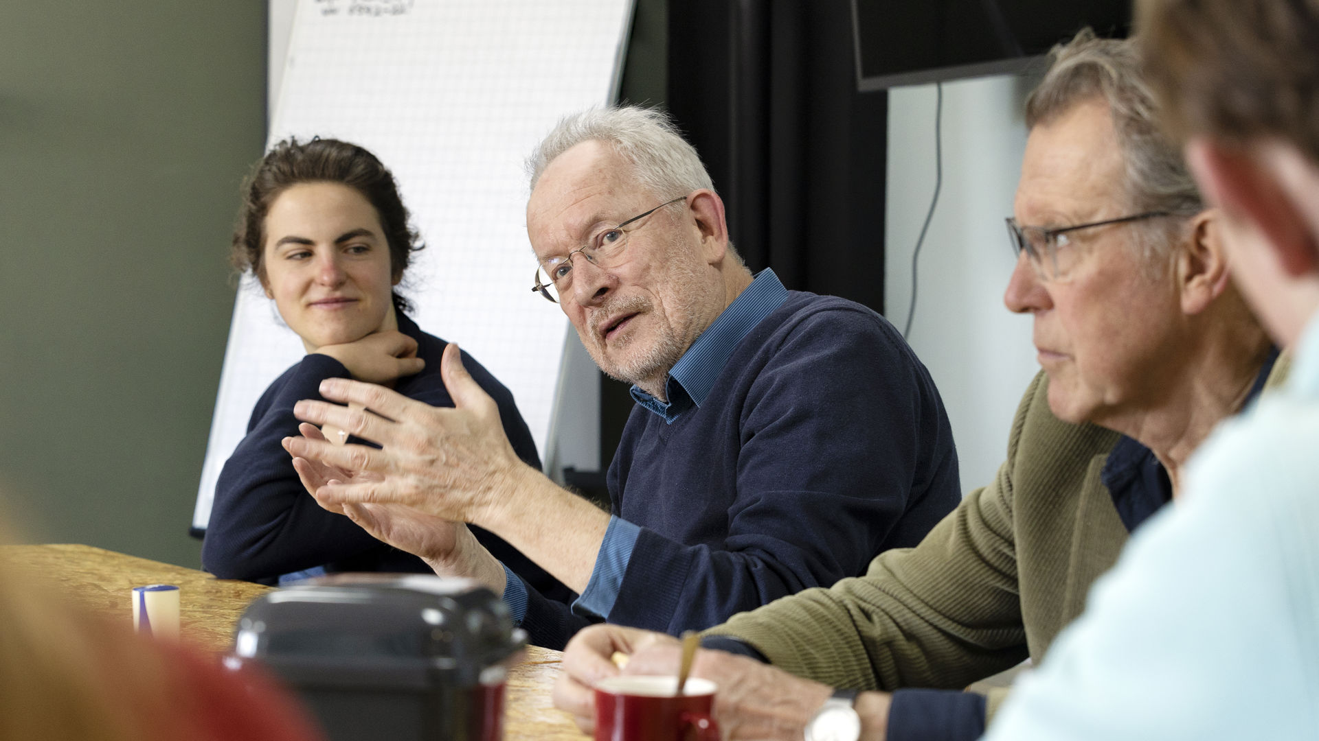 Huisarts Guus legt iets uit. Hij maakt handgebaren. Rondom hem zitten meer mensen aan tafel naar hem te luisteren.