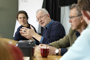 Huisarts Guus legt iets uit. Hij maakt handgebaren. Rondom hem zitten meer mensen aan tafel naar hem te luisteren.