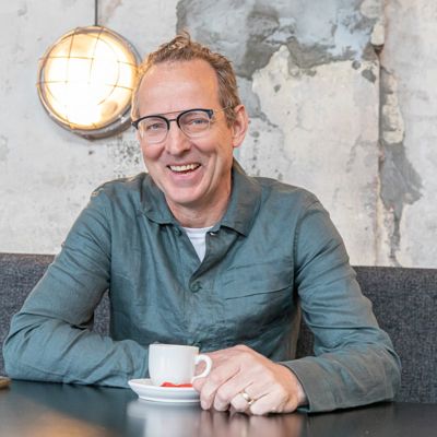 Miel zit achter een tafel met een kopje koffie voor zich. Hij draagt een grijze blouse en een bril met een half-montuur. Hij lacht in de camera.