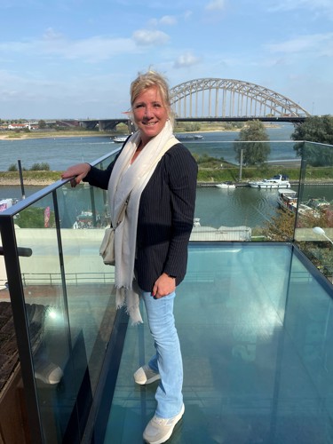 Nicolette staat op het glazen balkon van De Bastei. Ze draagt een spijkerbroek, een zwarte blazer en een witte sjaal. Ze heeft met een hand de reling vast.