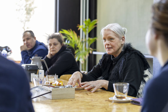 Een vrouw aan een tafel legt iets uit. Op de voor- en achtergrond zijn meer mensen die aan tafel zitten te zien.