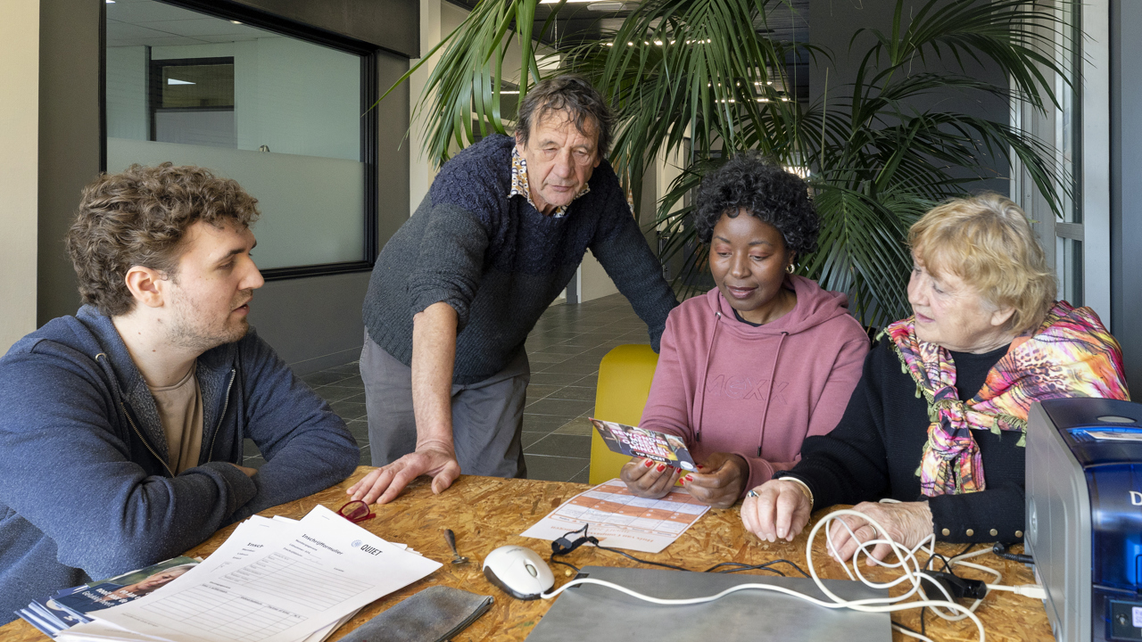 4 mensen zitten rond een tafel. Ze lezen een flyer. De tafel ligt vol met inschrijfspullen: een pasjesprinter, een laptop met muis, een etui, formulieren en flyers.